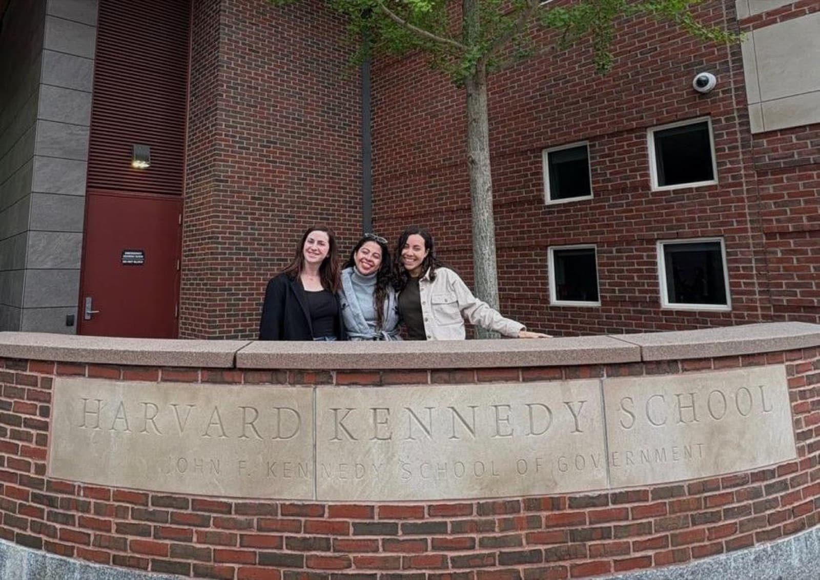 Daniela Schneider con dos compañeras frente al cartel de Harvard Kennedy School